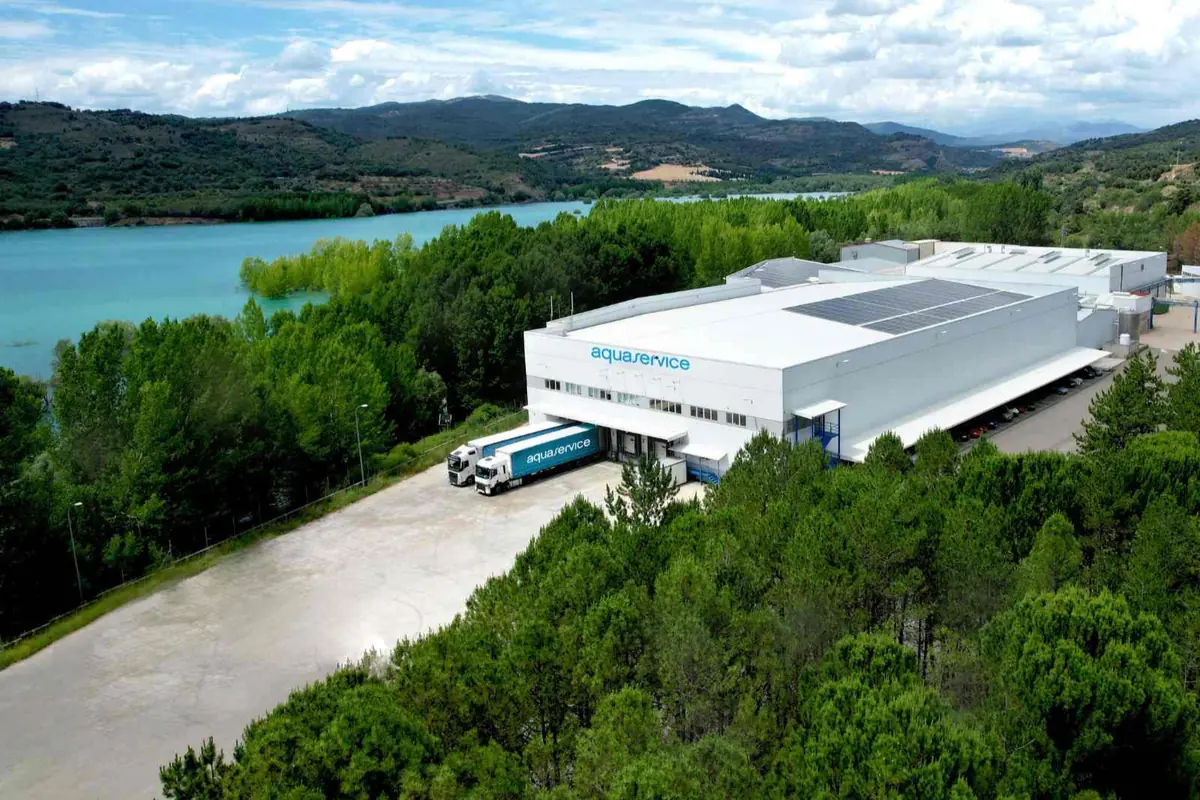 Vista aérea de una planta industrial de Aquaservice rodeada de árboles junto a un gran embalse de agua turquesa y montañas al fondo Vista aérea de una planta industrial de Aquaservice rodeada de árboles junto a un gran embalse de agua turquesa y montañas al fondo