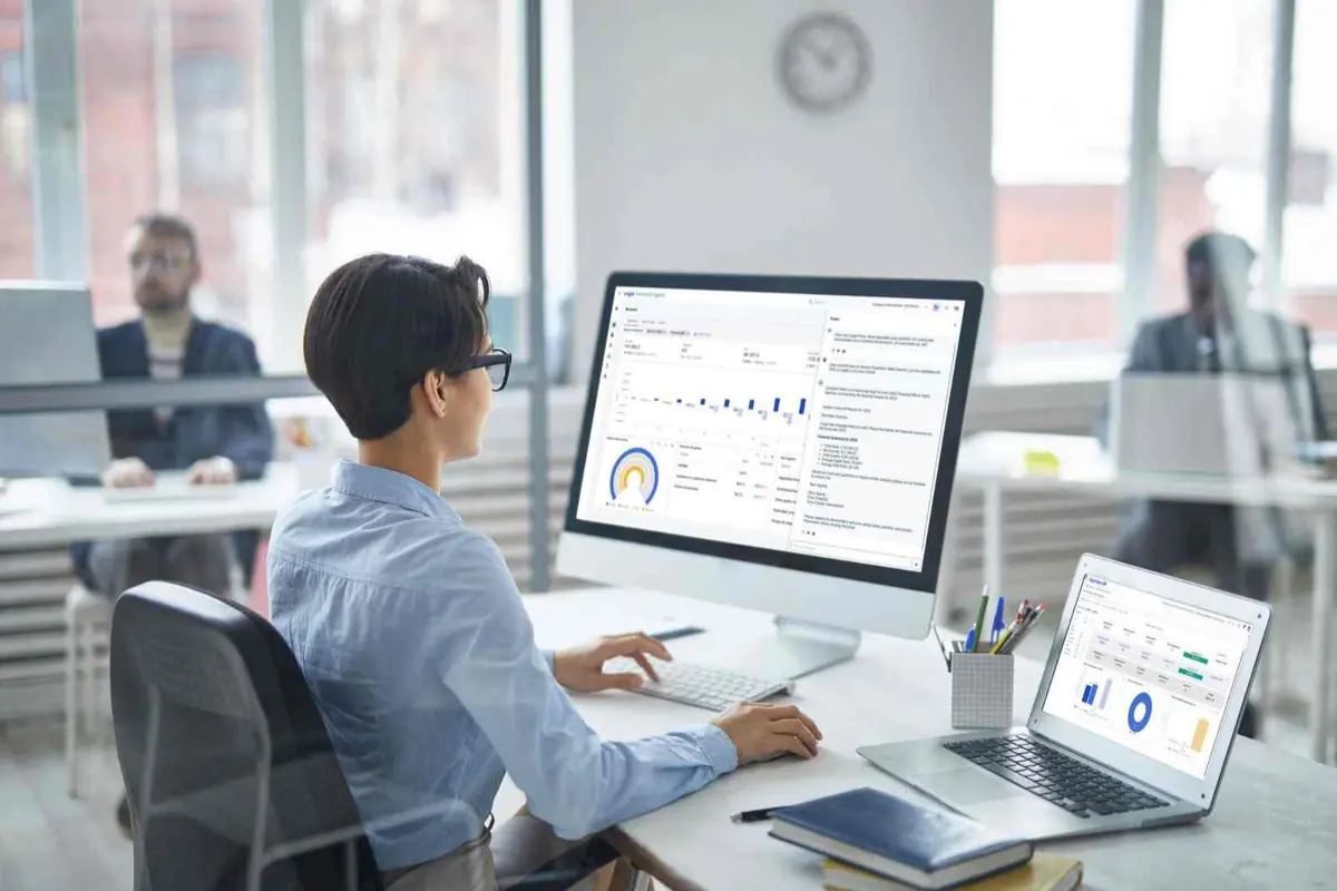 Persona trabajando en oficina moderna frente a un monitor y un portátil con paneles de análisis de datos y gráficos en pantalla
