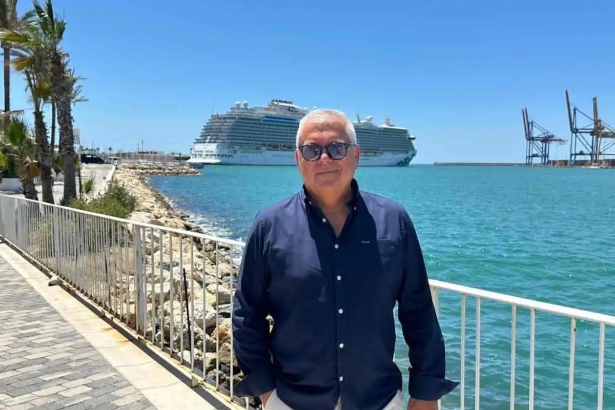 Hombre con gafas de sol posando junto al mar en un paseo marítimo con un gran crucero y grúas portuarias al fondo en un día soleado