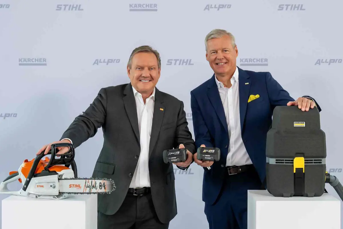 Dos ejecutivos de traje posan sonrientes mostrando baterías Allpro junto a una motosierra Stihl y un equipo de limpieza Kärcher sobre pedestales blancos Dos ejecutivos de traje posan sonrientes mostrando baterías Allpro junto a una motosierra Stihl y un equipo de limpieza Kärcher sobre pedestales blancos
