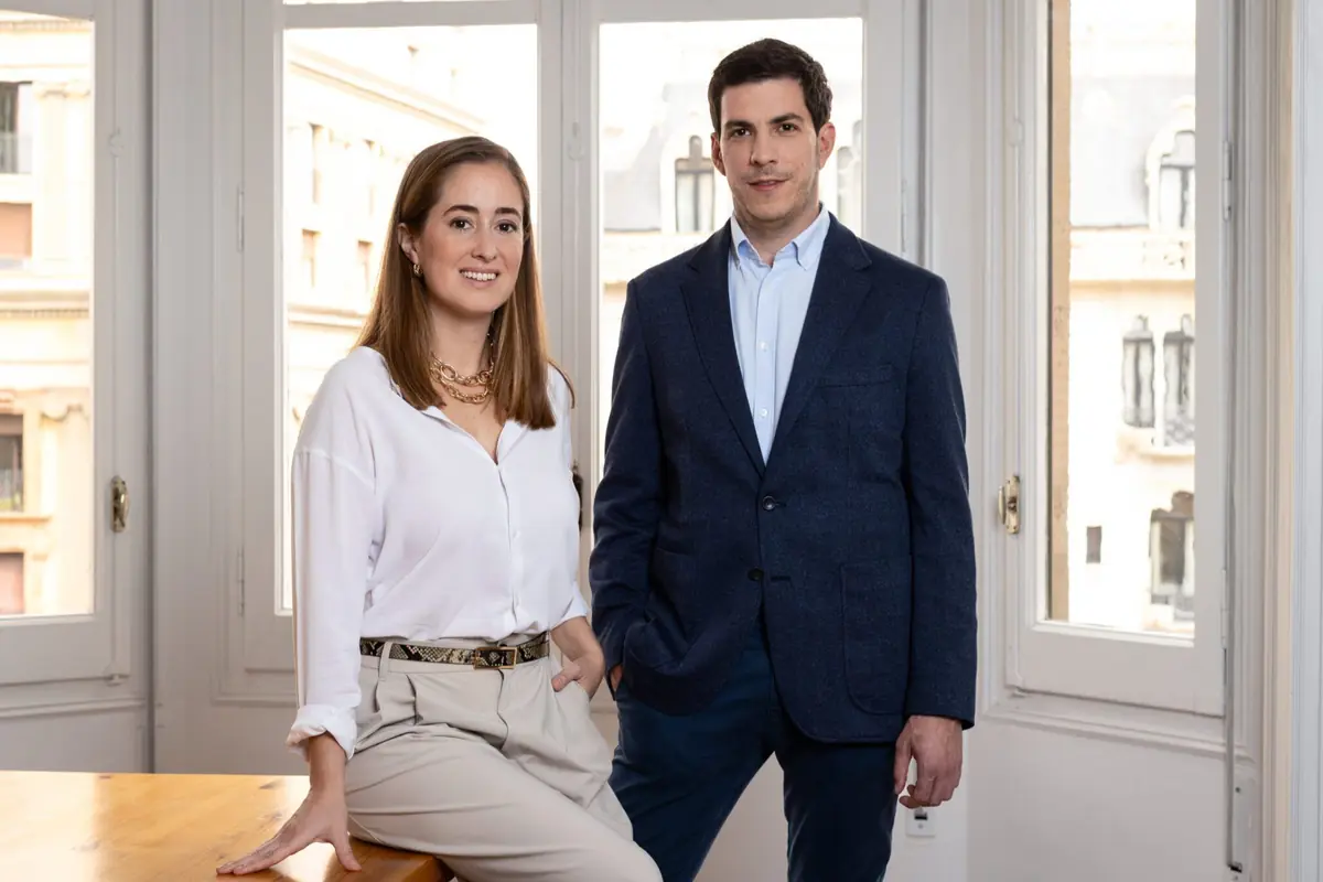 Dos personas jóvenes con ropa de oficina posan sonrientes en una sala luminosa con grandes ventanales y edificios de ciudad al fondo