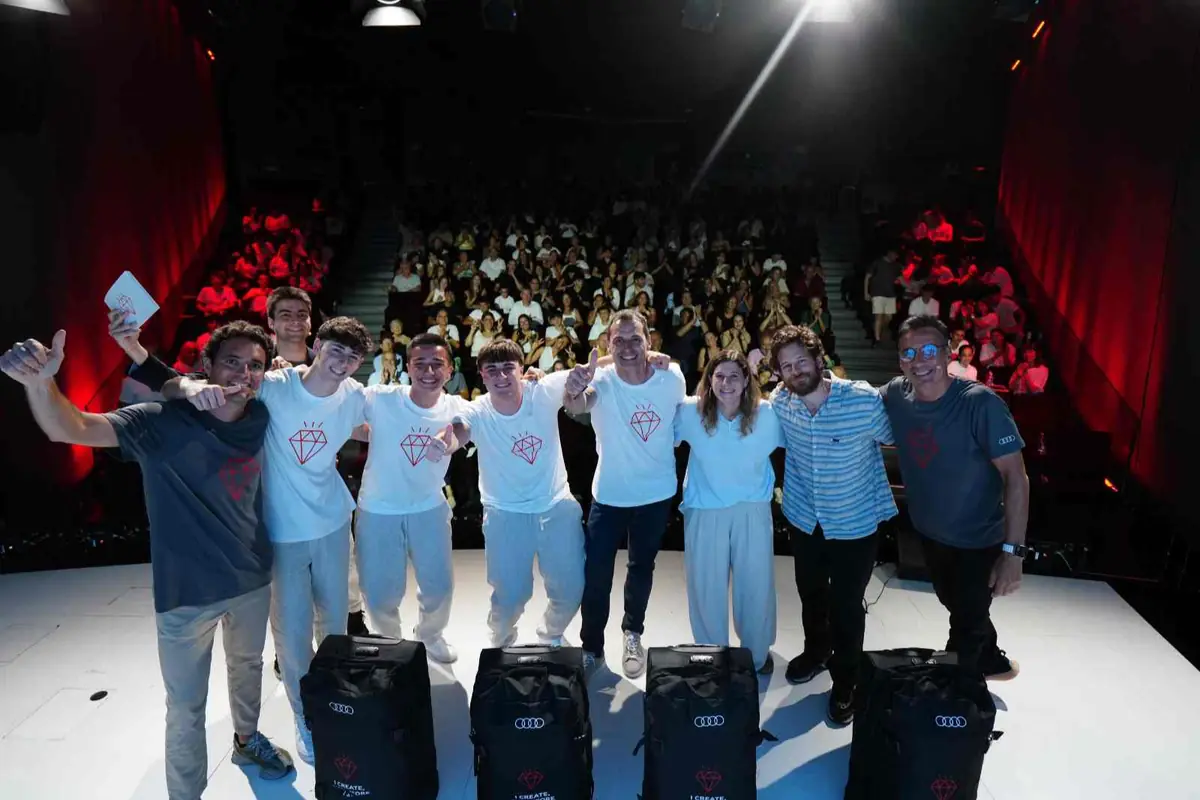 Grupo de jóvenes y adultos sonrientes posando en un escenario iluminado frente a un auditorio lleno durante un evento o presentación