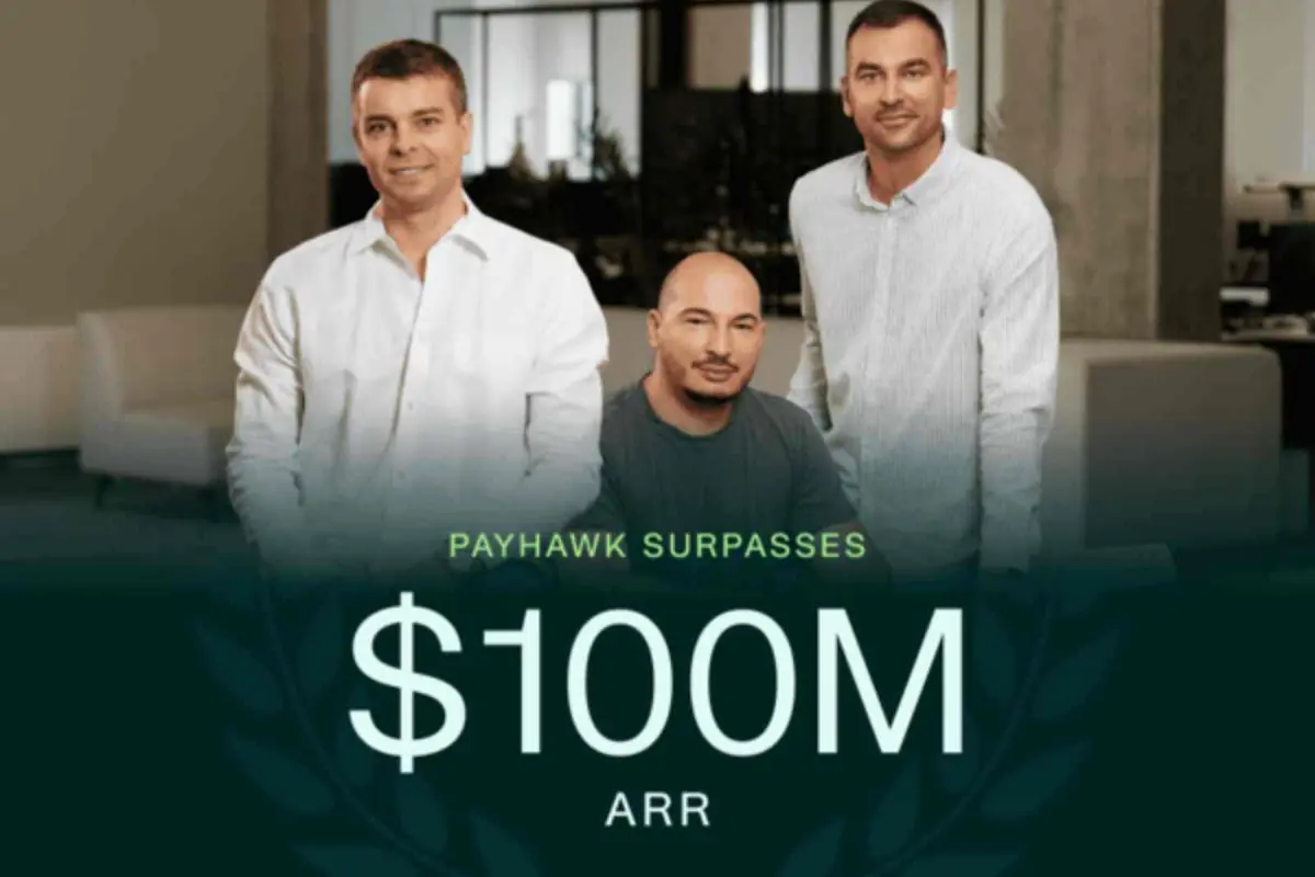 Tres hombres de negocios posan sonrientes en una oficina detrás de un gran texto que anuncia que Payhawk supera los 100 millones de dólares de ingresos recurrentes anuales