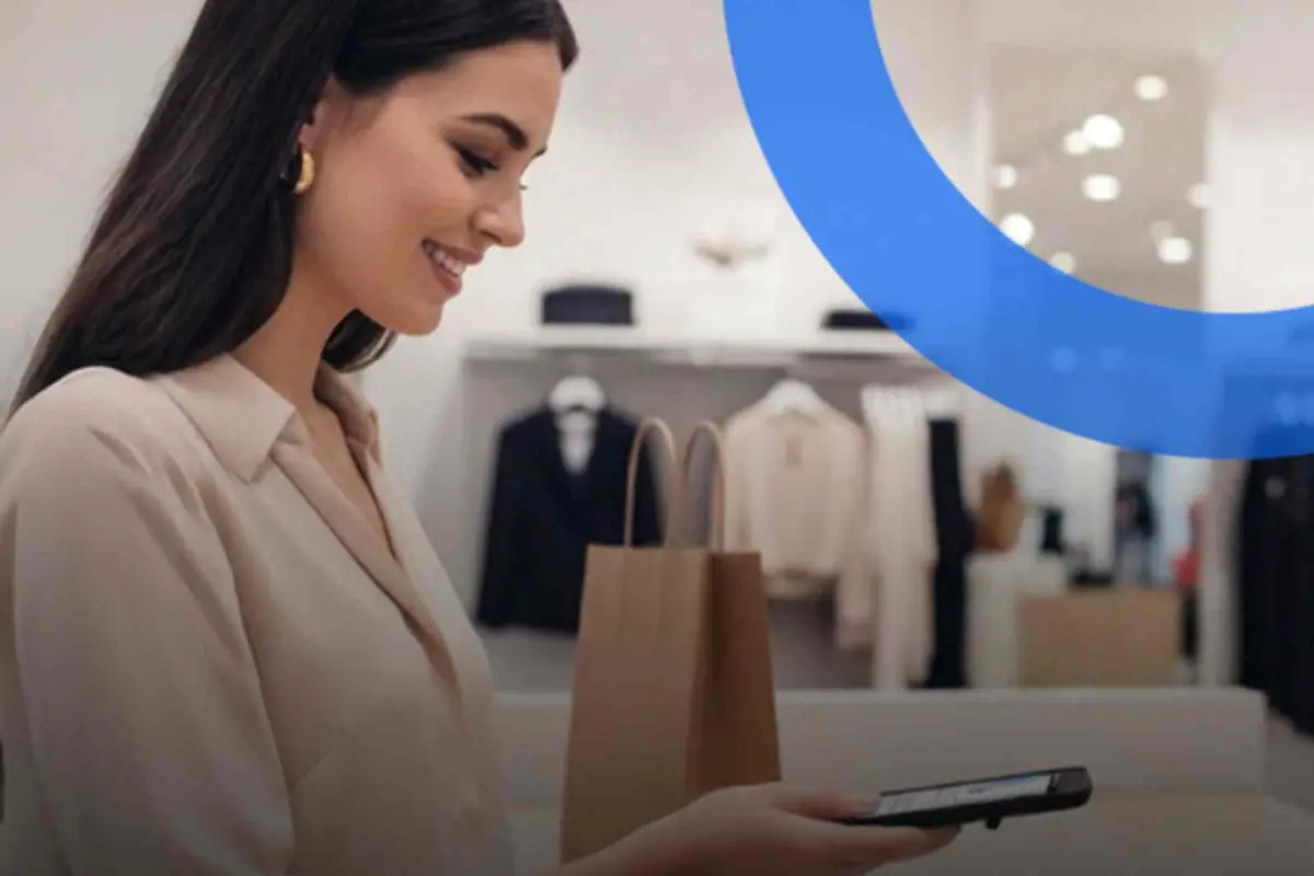 Mujer joven sonriendo mientras revisa su teléfono móvil en una tienda de ropa con una bolsa de compras sobre el mostrador