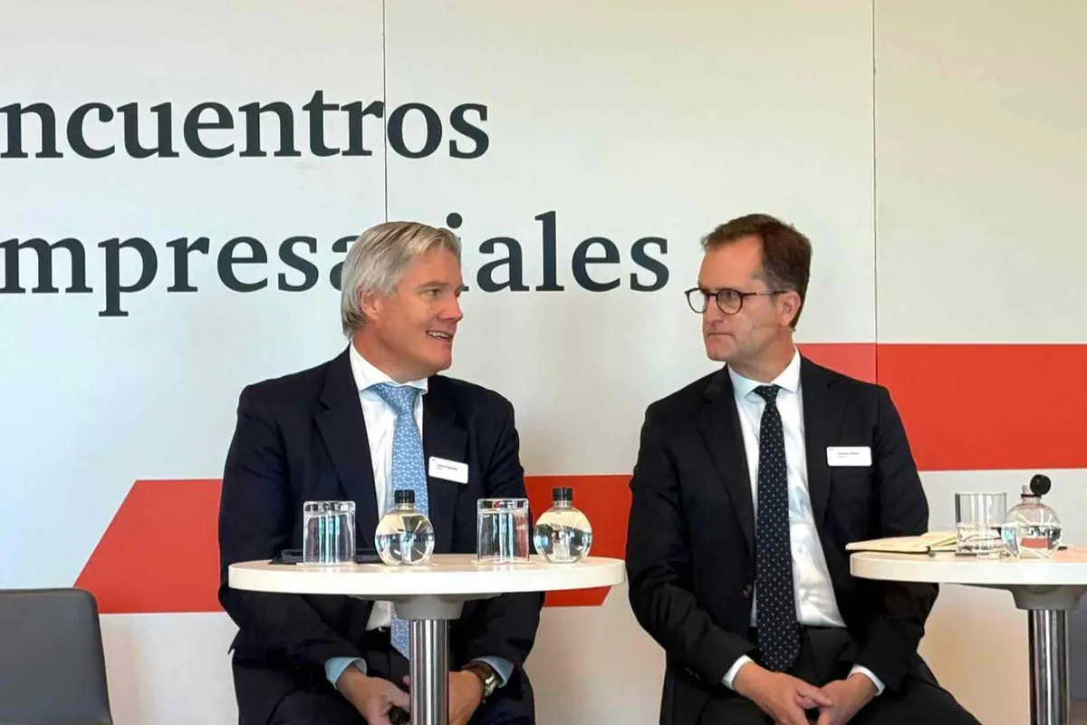 Dos hombres con traje conversan sentados en una mesa redonda durante un encuentro empresarial con vasos y botellas de agua sobre la mesa y un cartel de fondo con el texto encuentros empresariales Dos hombres con traje conversan sentados en una mesa redonda durante un encuentro empresarial con vasos y botellas de agua sobre la mesa y un cartel de fondo con el texto encuentros empresariales