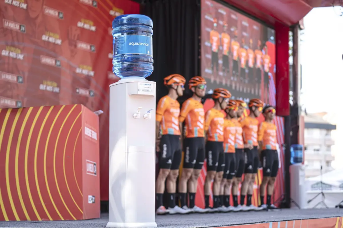 Dispensador de agua en primer plano con un equipo de ciclistas uniformados de naranja alineados en un escenario de presentación deportiva al fondo