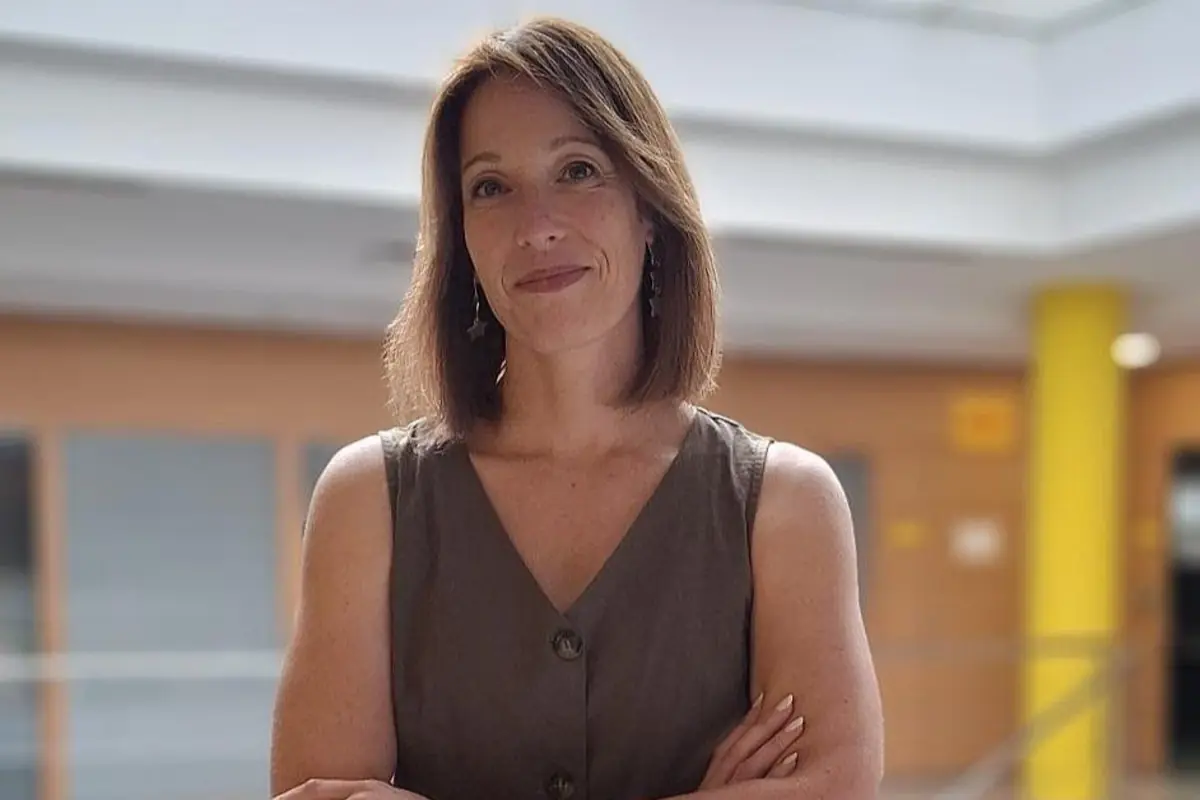 Mujer de mediana edad con vestido sin mangas de color marrón posando con los brazos cruzados en un interior luminoso con columnas amarillas de fondo