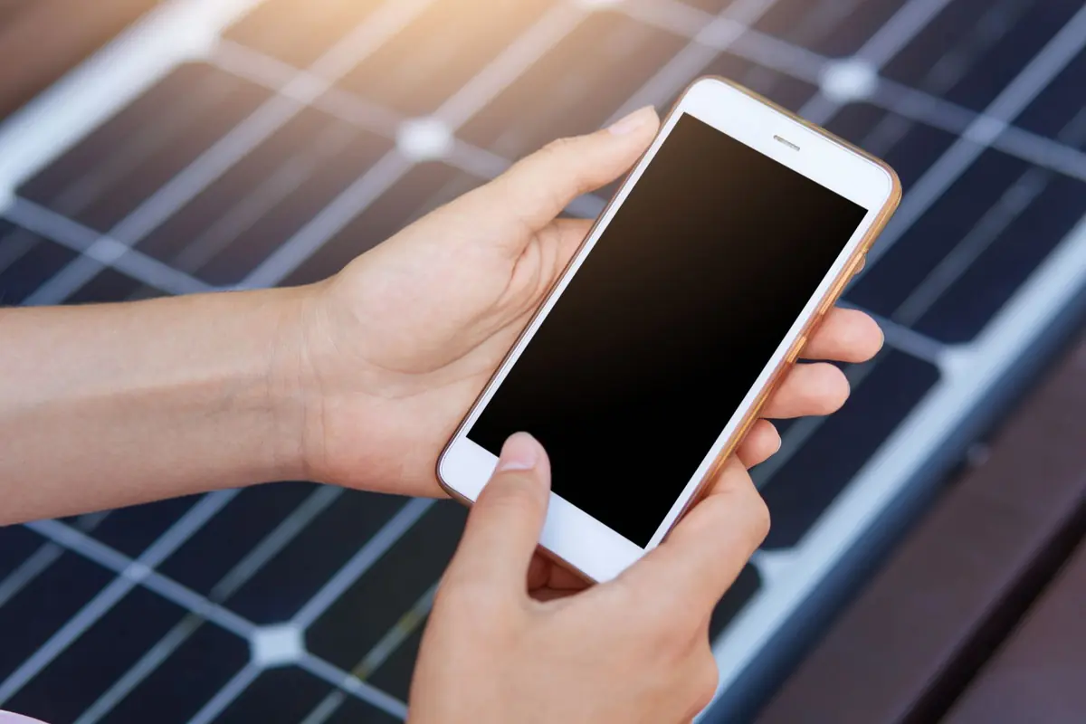 Manos sosteniendo un teléfono móvil con pantalla apagada frente a paneles solares