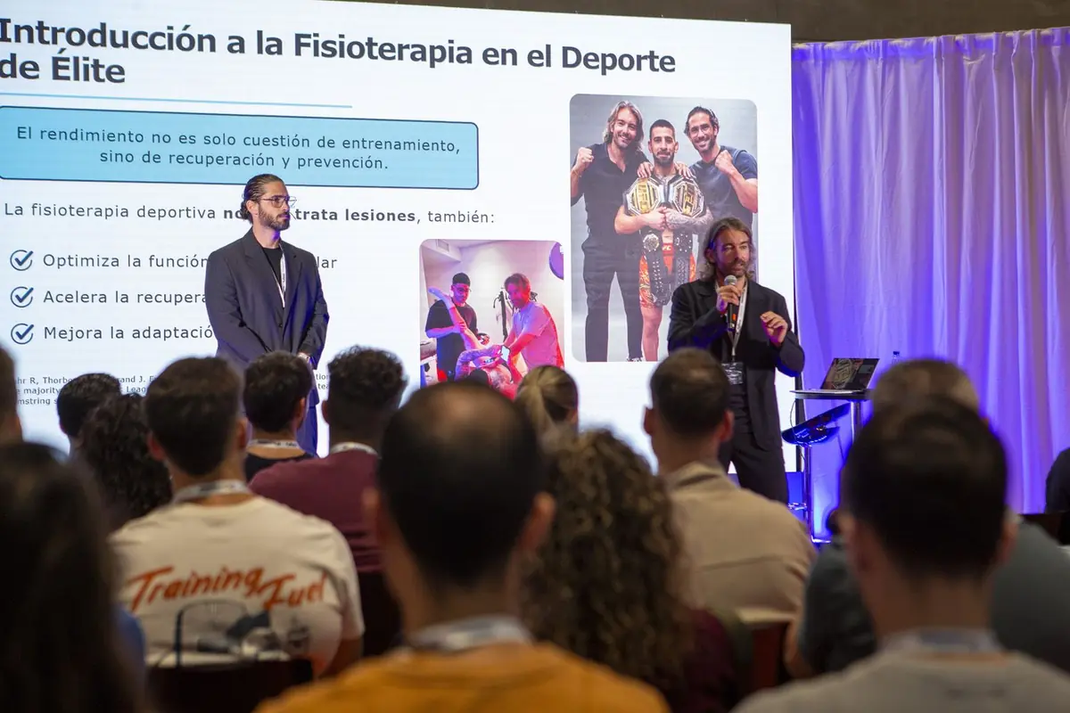 Ponentes impartiendo una conferencia sobre fisioterapia en el deporte de élite ante un auditorio lleno, con una gran pantalla proyectando diapositivas y fotografías de deportistas profesionales
