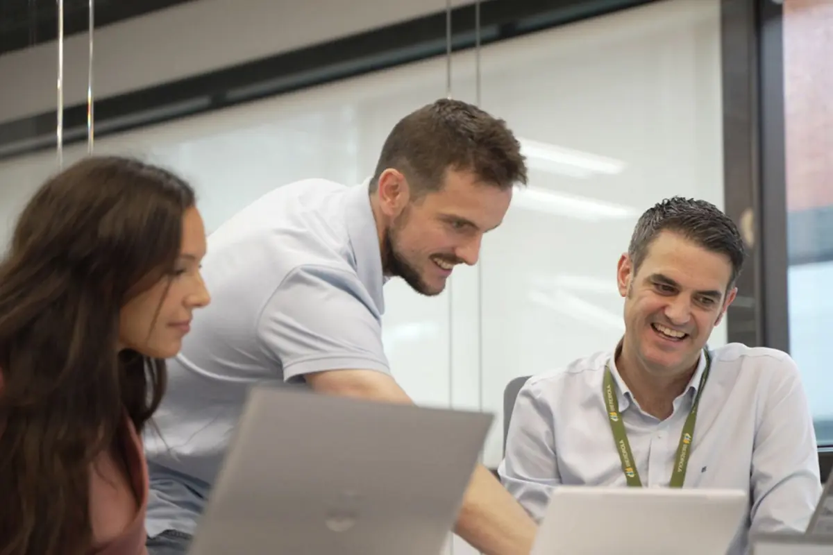 Tres personas trabajando juntas y sonriendo mientras revisan información en sus portátiles en una sala de reuniones moderna