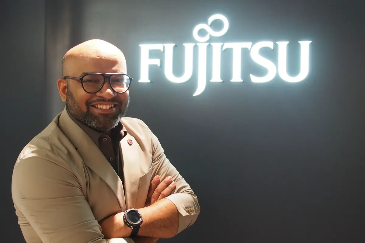 Hombre con gafas y barba sonriendo con los brazos cruzados frente a un letrero iluminado de Fujitsu