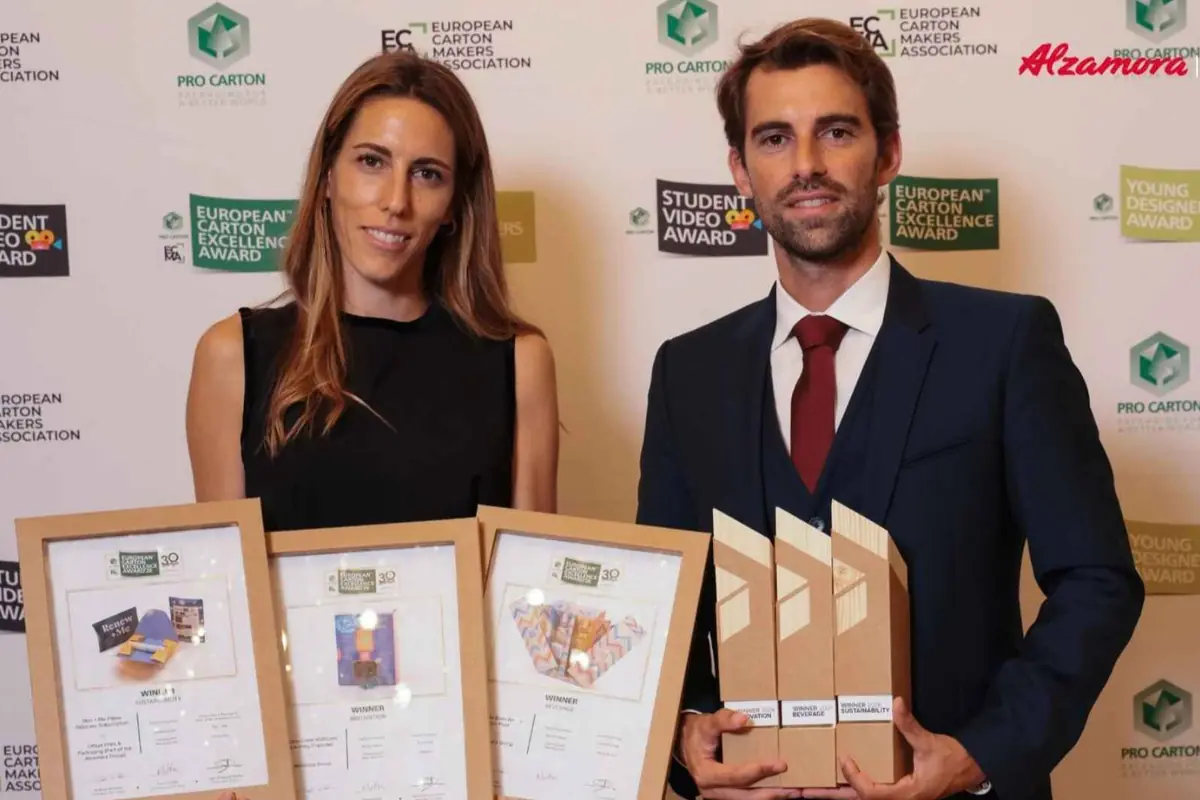Dos personas posan ante un photocall de premios de diseño de envases sosteniendo varios diplomas y trofeos de cartón