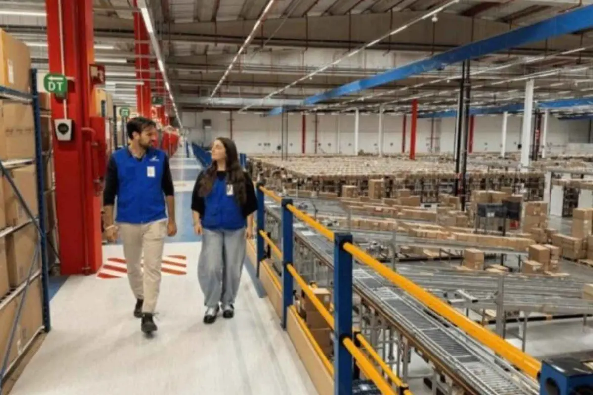 Dos empleados con chalecos azules caminan por una pasarela elevada en un gran almacén logístico lleno de estanterías y cajas de cartón