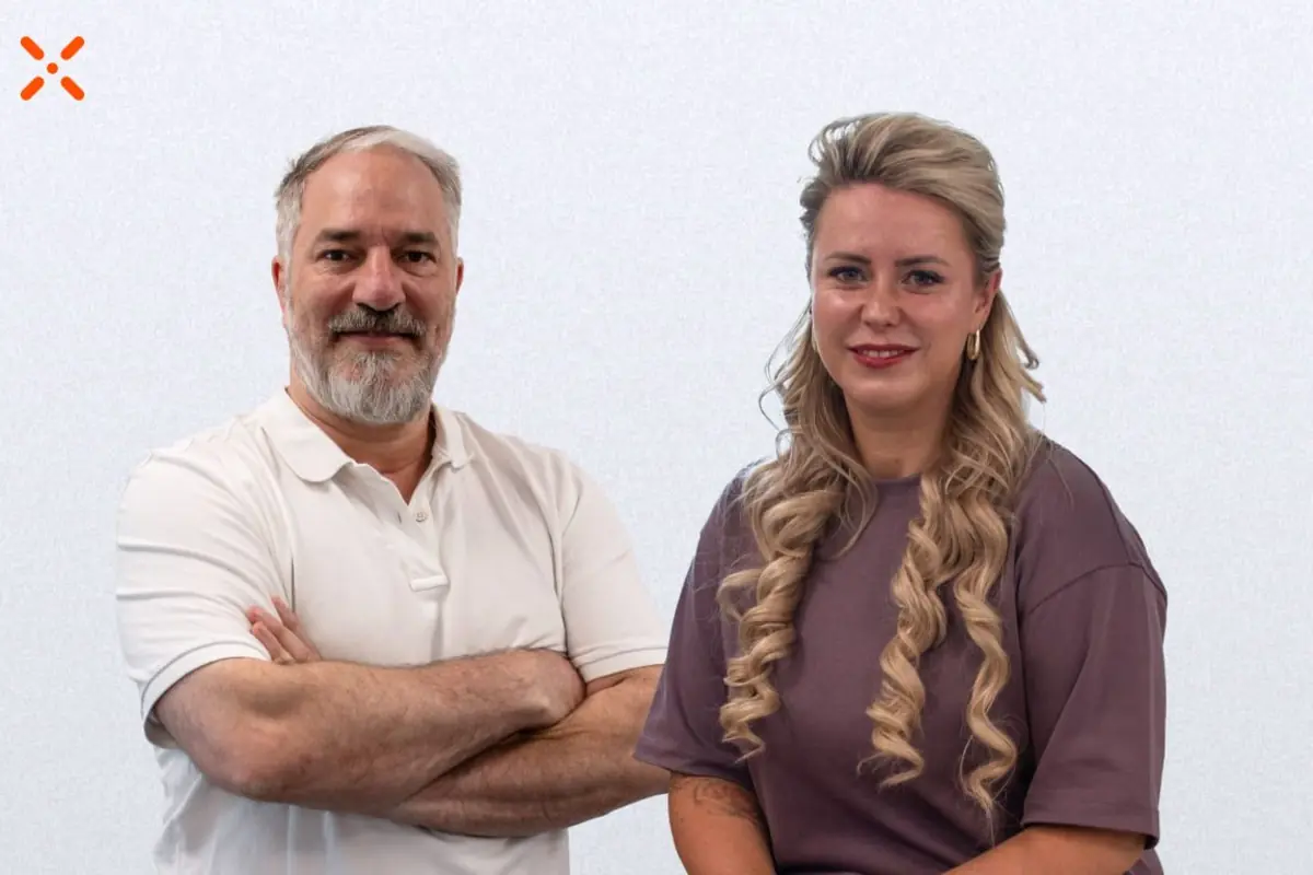 Hombre de mediana edad con barba canosa y brazos cruzados junto a mujer rubia de pelo largo rizado ambos sonriendo frente a un fondo claro