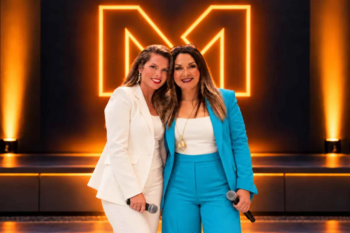 Dos mujeres sonrientes con micrófonos posan juntas en un escenario iluminado con luces naranjas y un gran logo con forma de letra M al fondo
