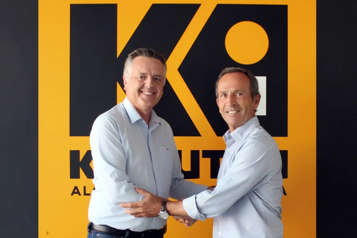 Dos hombres con camisa clara se dan la mano y sonríen frente a un gran logotipo amarillo y negro en una pared