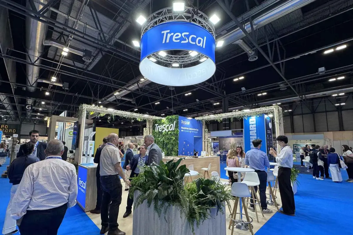 Stand de Trescal en una feria profesional con personas conversando, plantas decorativas y mesas altas sobre moqueta azul en un recinto interior amplio