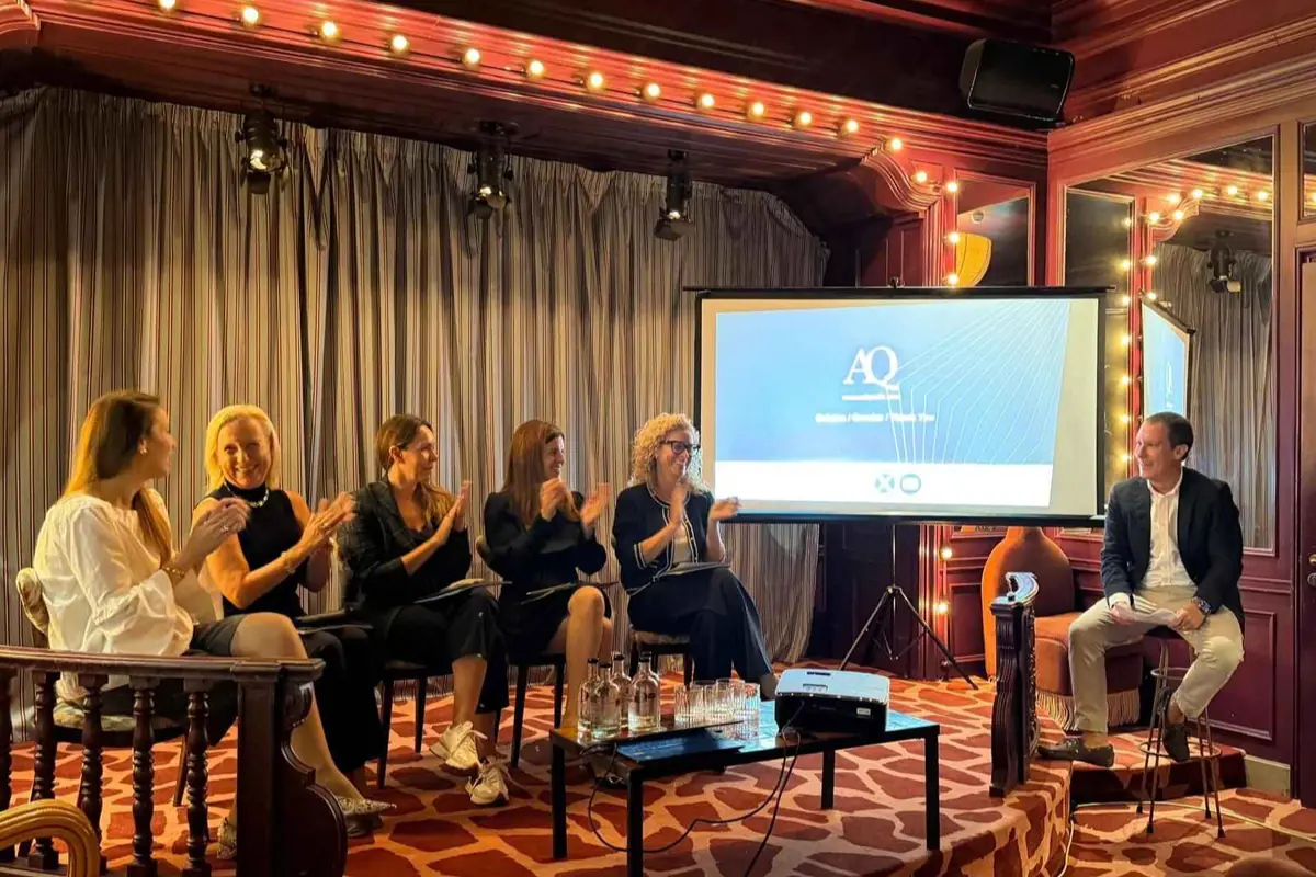 Panel de seis personas en un salón elegante aplaudiendo mientras un ponente se sienta junto a una pantalla de presentación con el logotipo AQ