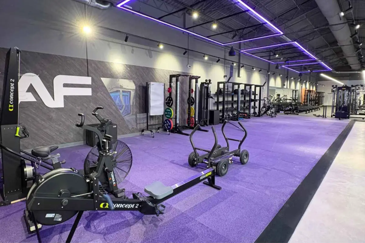 Gimnasio moderno con piso morado máquinas de remo y cardio en primer plano y área amplia de entrenamiento con pesas y racks al fondo iluminación de tiras LED púrpuras en el techo