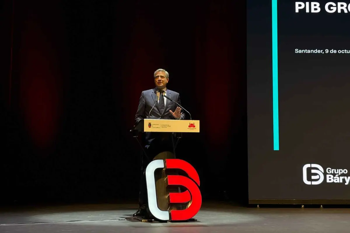 Hombre con traje dando un discurso en un auditorio desde un atril iluminado con un gran logotipo rojo y blanco frente a una pantalla de presentación oscura