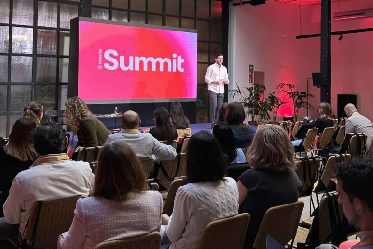 Hombre dando una charla en un escenario con pantalla grande que muestra la palabra Summit frente a un público sentado en una sala de conferencias moderna