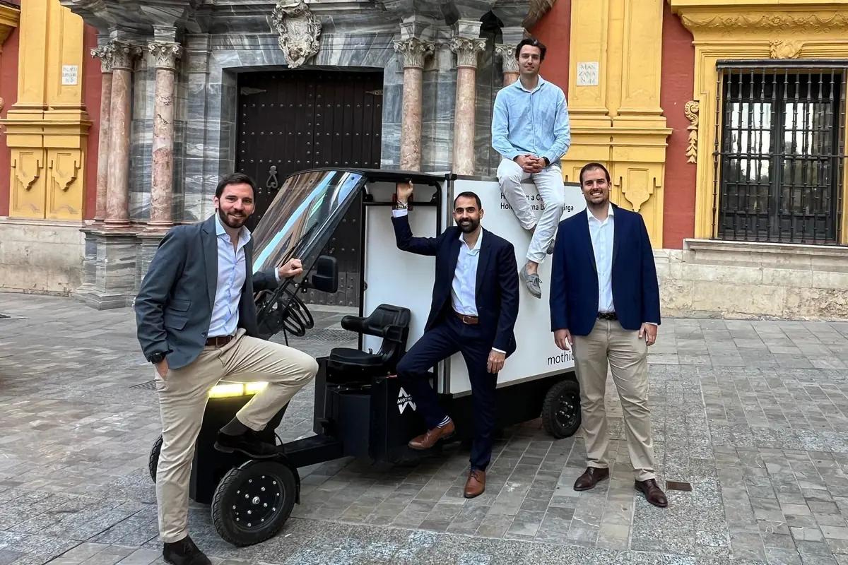 Cuatro hombres con ropa formal posan sonrientes alrededor de un pequeño vehículo eléctrico de reparto en una plaza con edificio histórico de fondo