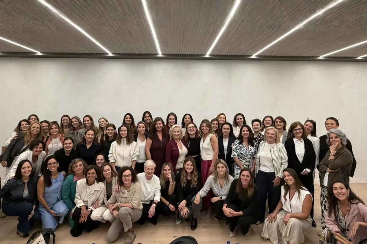 Grupo numeroso de mujeres de distintas edades posando sonrientes en el interior de una sala moderna con techo de luces lineales