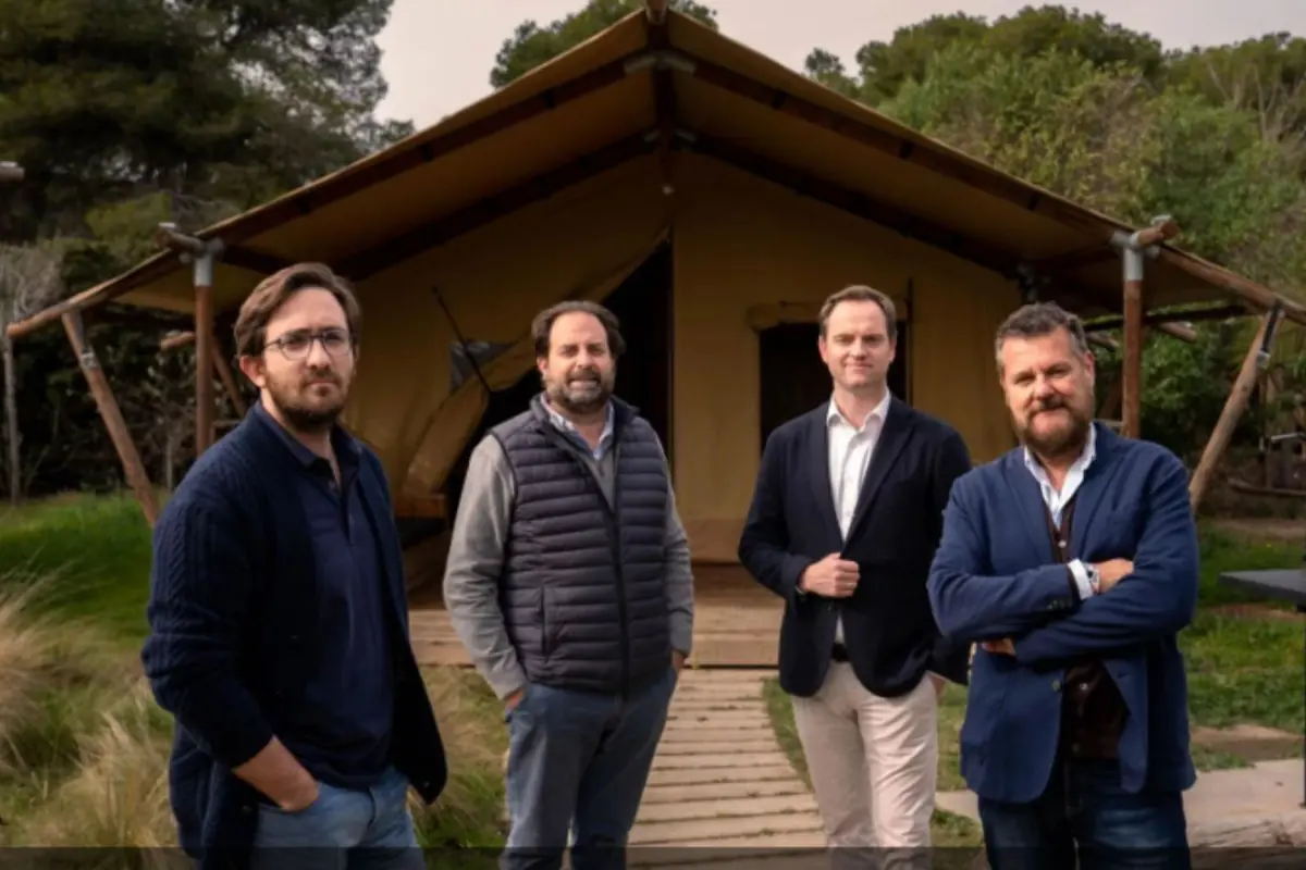 Cuatro hombres posan frente a una gran tienda tipo glamping en un entorno natural con árboles y césped