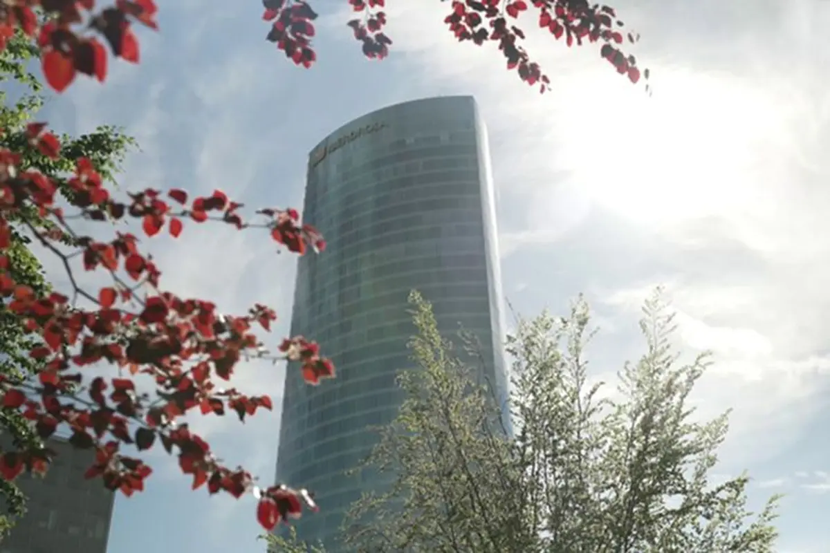 Edificio moderno de cristal visto desde abajo con ramas de árboles y hojas rojas en primer plano bajo un cielo soleado