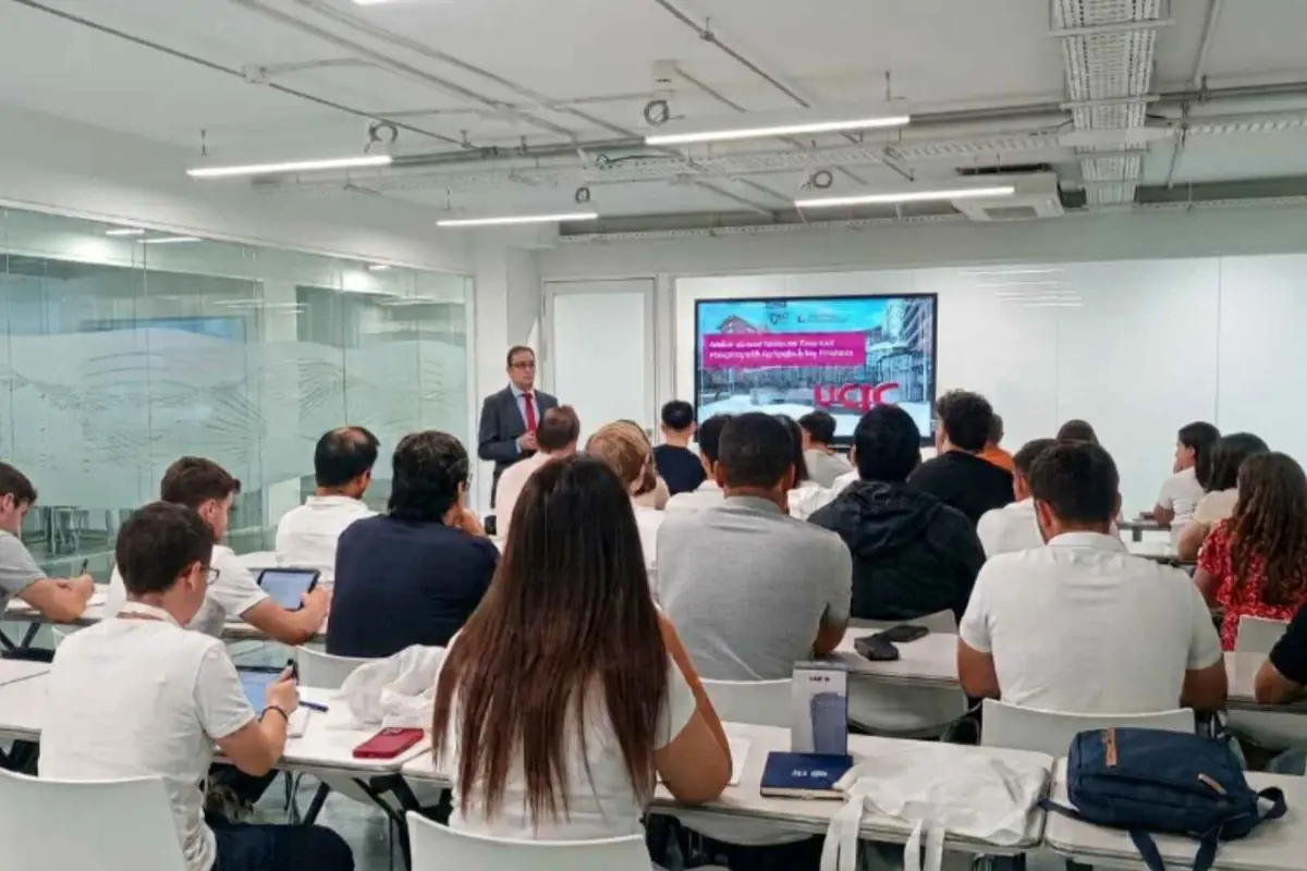 Profesor dando una presentación en una sala moderna mientras un grupo de estudiantes atiende sentado frente a una pantalla grande con diapositivas