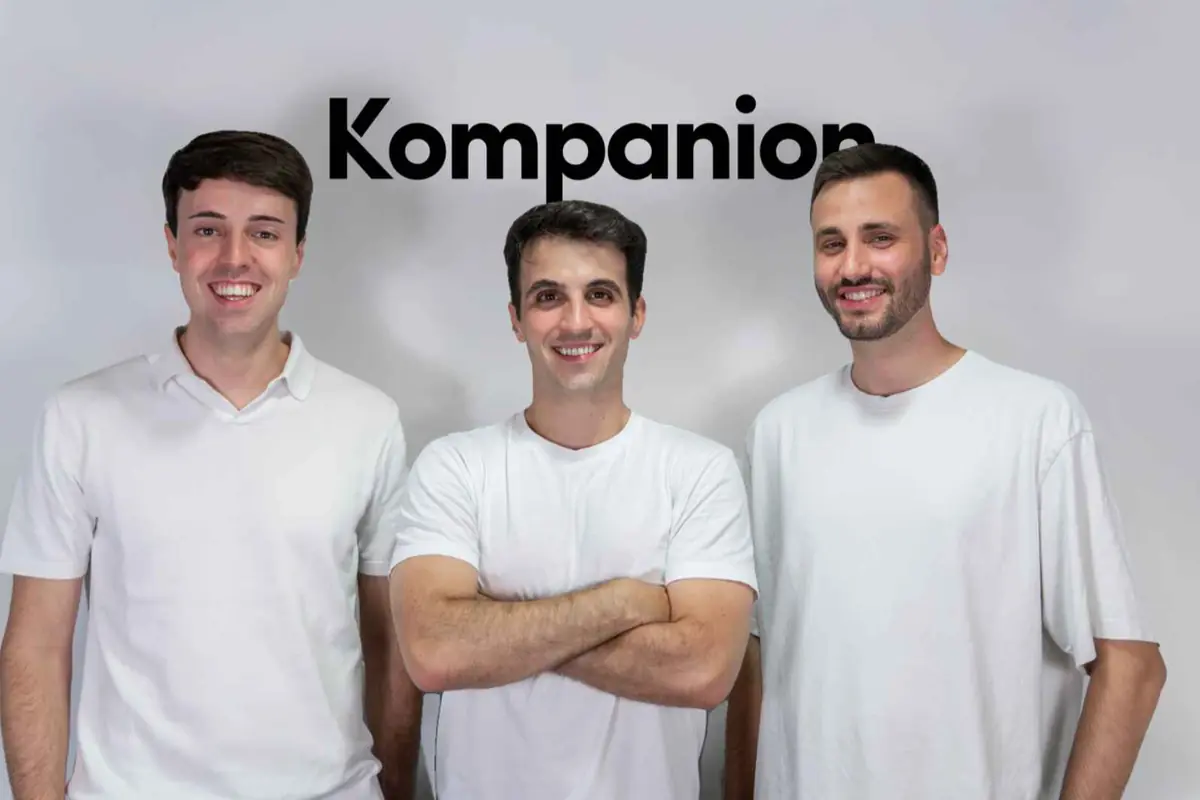 Tres hombres jóvenes con camisetas blancas posan sonriendo frente a un fondo claro con el logotipo de Kompanion encima de ellos Tres hombres jóvenes con camisetas blancas posan sonriendo frente a un fondo claro con el logotipo de Kompanion encima de ellos