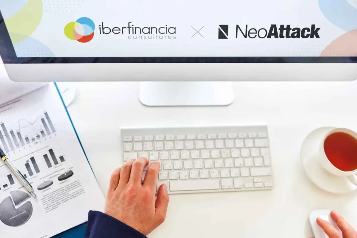 Escritorio de oficina con ordenador que muestra la colaboración entre Iberfinancia Consultores y NeoAttack junto a teclado gráficos financieros y una taza de té