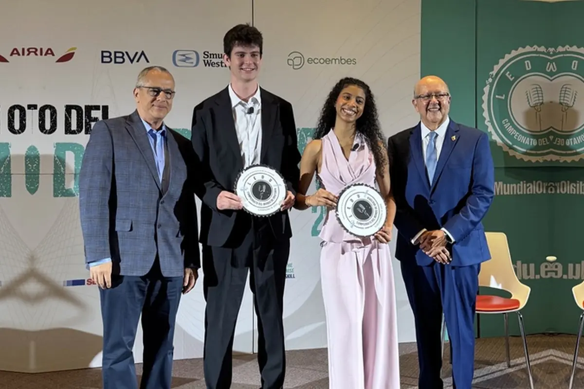 Cuatro personas posan de pie en un escenario de premiación dos jóvenes en el centro sostienen placas circulares mientras dos hombres mayores con traje los acompañan delante de un fondo con logotipos de patrocinadores Cuatro personas posan de pie en un escenario de premiación dos jóvenes en el centro sostienen placas circulares mientras dos hombres mayores con traje los acompañan delante de un fondo con logotipos de patrocinadores