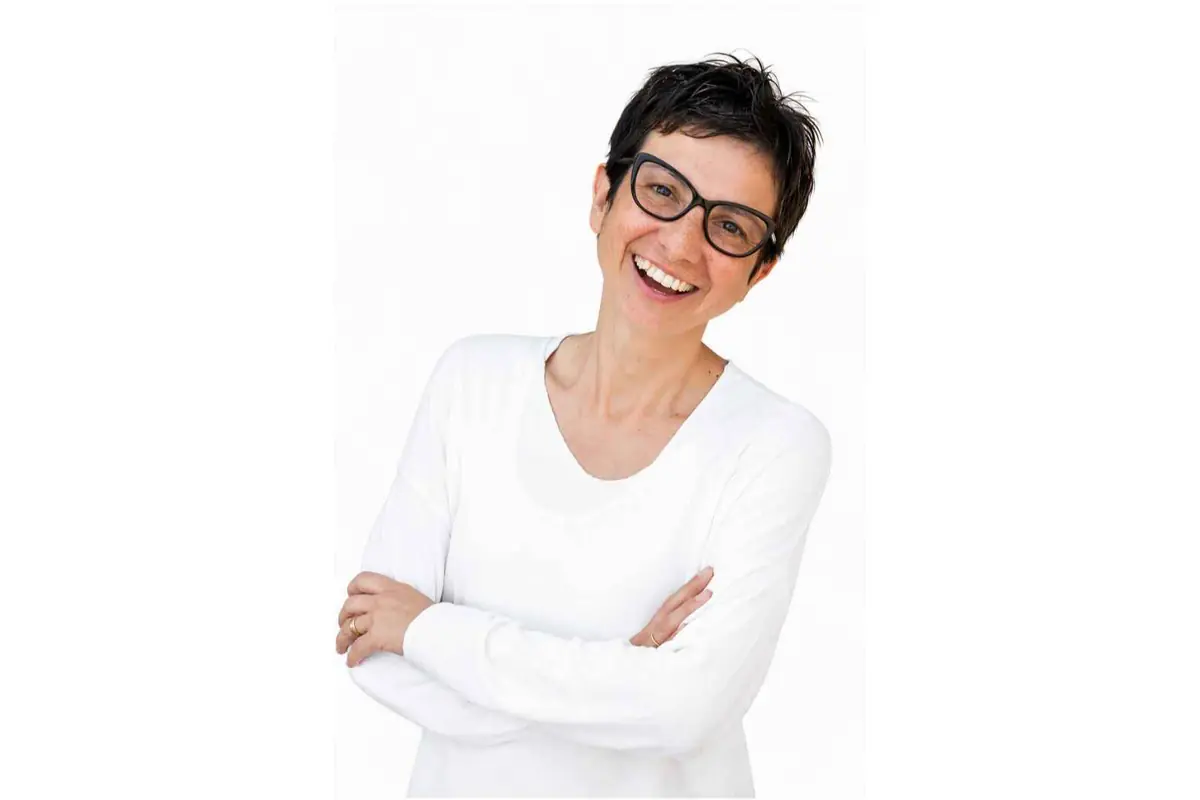 Mujer sonriente con gafas negras y camiseta blanca posando con los brazos cruzados sobre fondo blanco