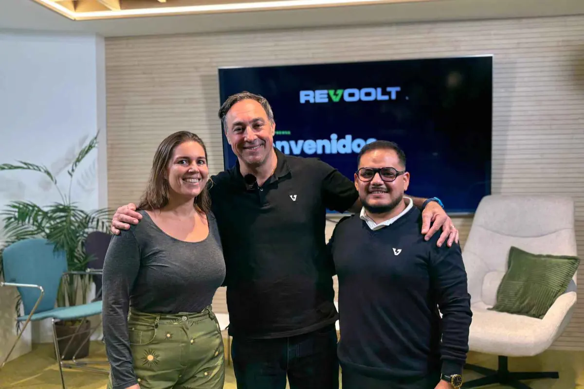Tres personas sonriendo posan abrazadas en una sala moderna frente a una pantalla con el logo de Revoolt y la palabra Bienvenidos