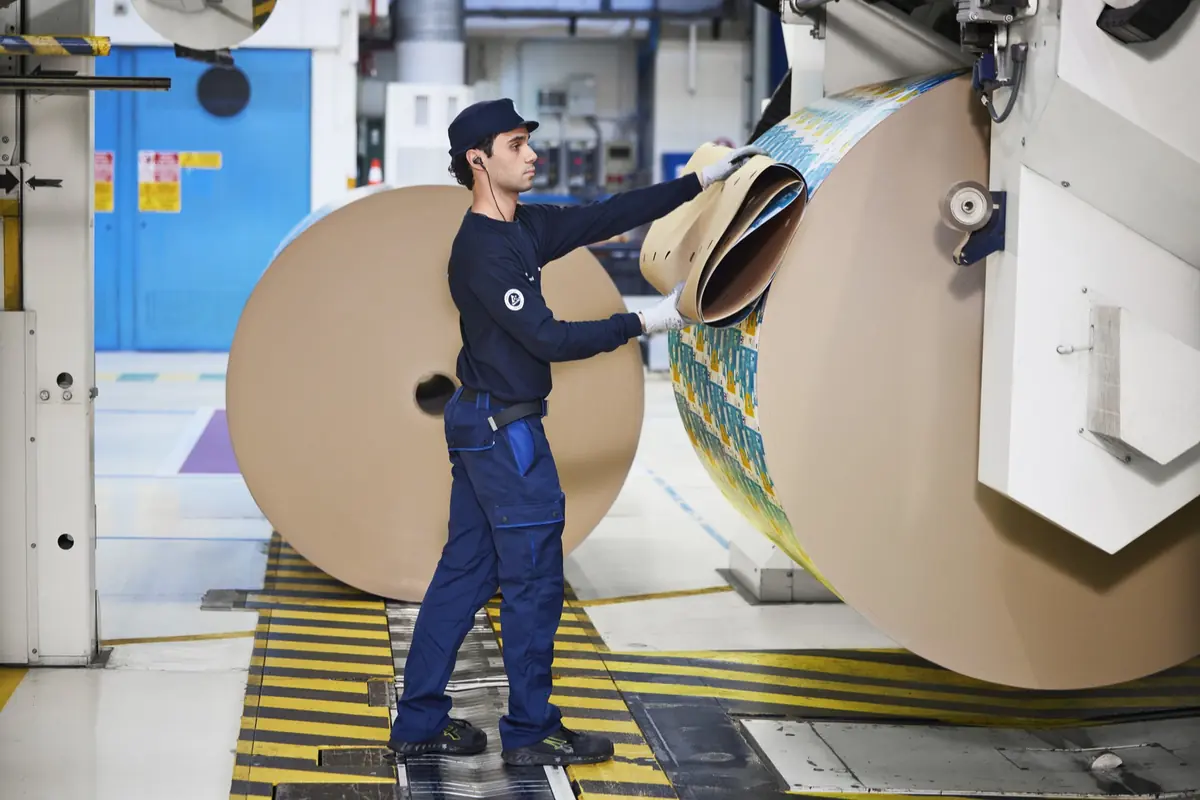 Trabajador industrial manipulando una gran bobina de papel en una máquina de impresión dentro de una fábrica Trabajador industrial manipulando una gran bobina de papel en una máquina de impresión dentro de una fábrica