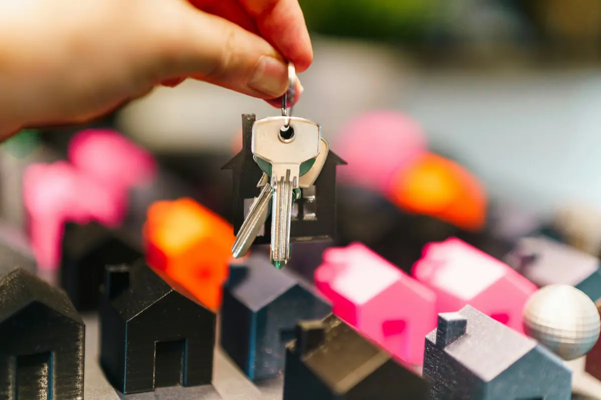 Mano sosteniendo llavero con llaves de casa sobre maqueta de pequeñas viviendas de colores