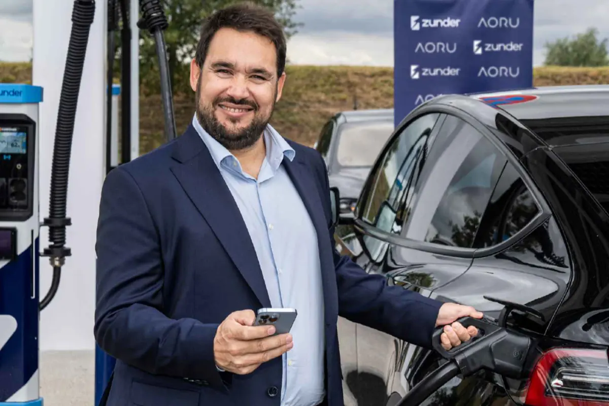 Hombre sonriente con traje cargando un coche eléctrico negro mientras sostiene un teléfono móvil en una estación de carga Hombre sonriente con traje cargando un coche eléctrico negro mientras sostiene un teléfono móvil en una estación de carga