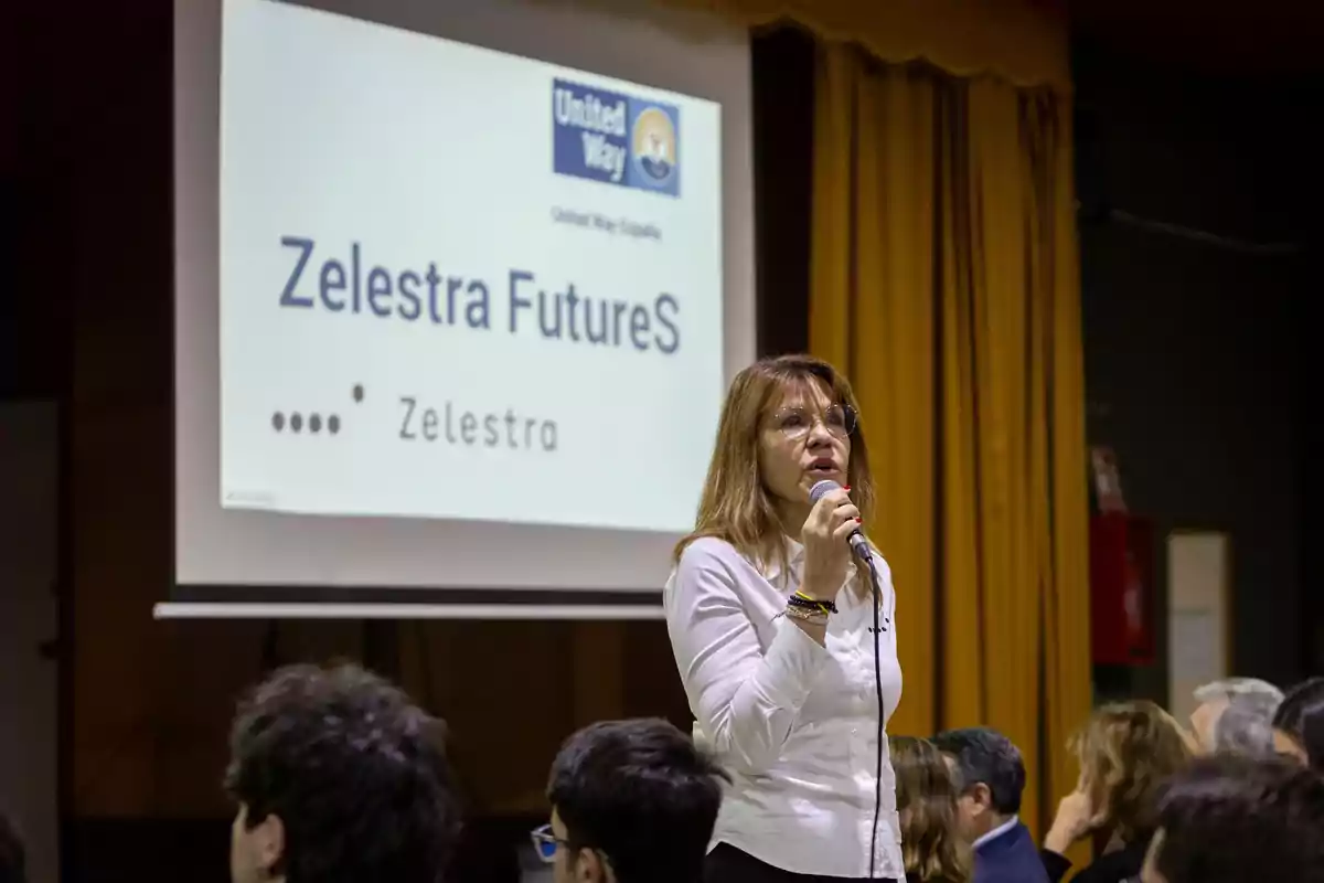 Mujer hablando con micrófono frente a una audiencia, con una pantalla de presentación al fondo que muestra el texto Zelestra FutureS y el logo de United Way