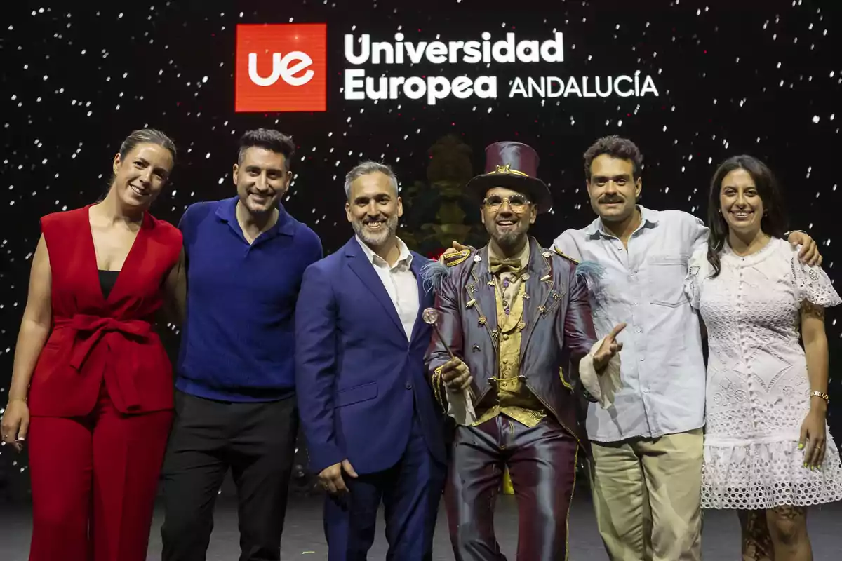 Seis personas posan sonrientes en un escenario frente a un fondo con el logo de la Universidad Europea de Andalucía, una de ellas lleva un colorido disfraz y sombrero de copa.