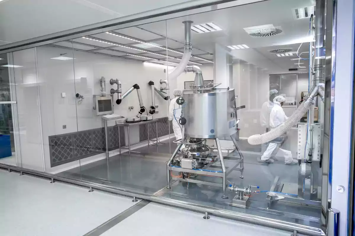 Una sala limpia de laboratorio con equipos de acero inoxidable y personas con trajes protectores trabajando en su interior.