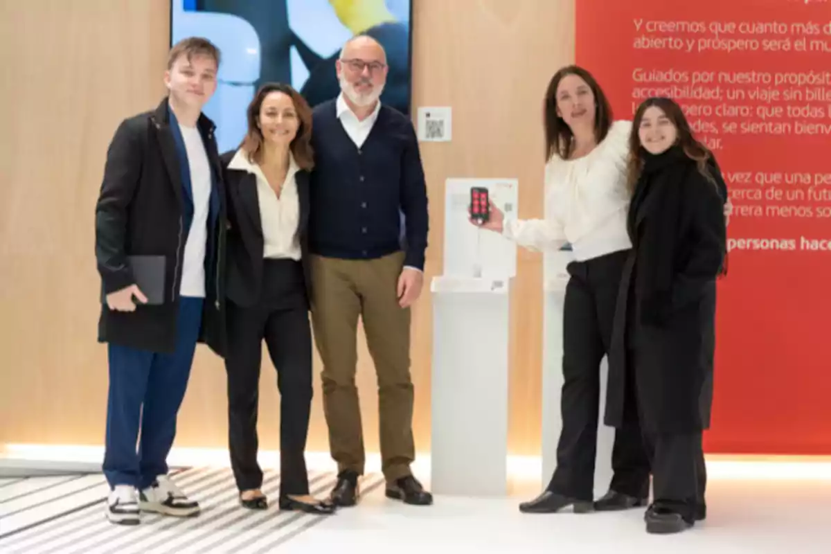 Un grupo de cinco personas posando juntas en un entorno moderno y bien iluminado, con una de ellas sosteniendo un dispositivo móvil frente a un panel informativo rojo.