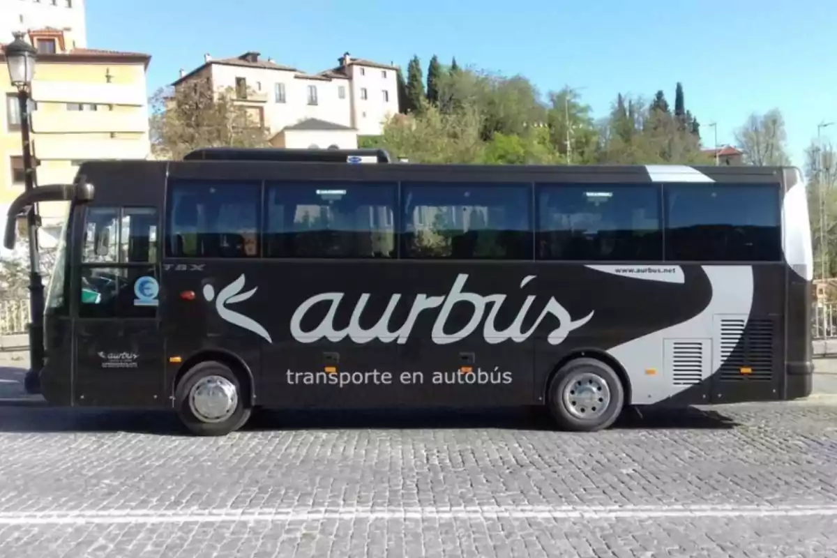 Autobús negro de la empresa "Aurbús" estacionado en una calle empedrada con edificios y árboles al fondo.