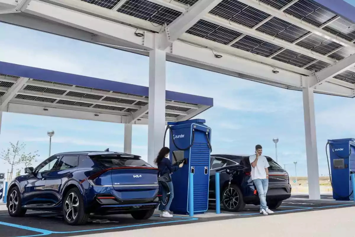 Dos autos eléctricos están estacionados y cargando en una estación de carga bajo un techo con paneles solares, mientras dos personas interactúan con los cargadores.