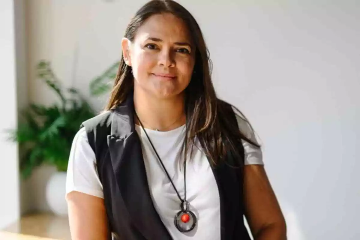 Una persona con cabello largo y oscuro, vestida con camiseta blanca y chaleco negro, lleva un collar con un colgante rojo y está de pie frente a una planta en un entorno bien iluminado.