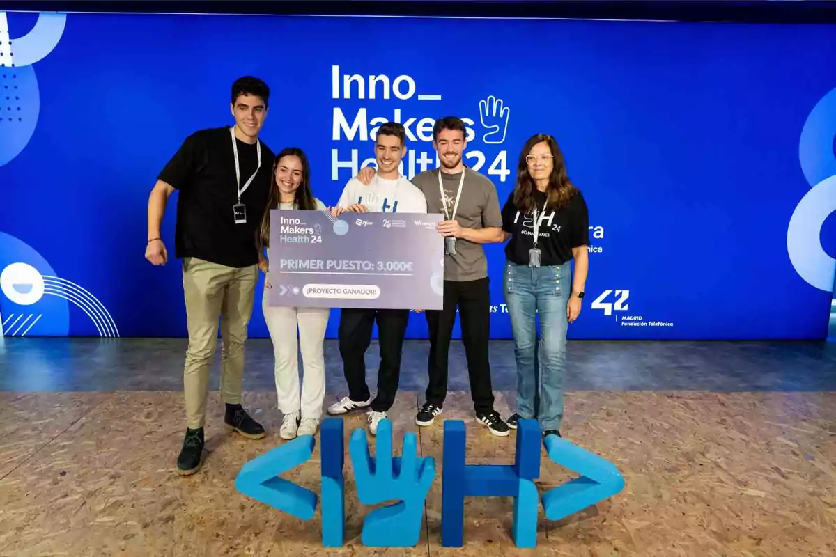 Un grupo de cinco personas sonríe mientras sostiene un cheque grande que indica que han ganado el primer puesto en un evento llamado Inno Makers Health 24, con un fondo azul que muestra el nombre del evento y un logotipo en el suelo.