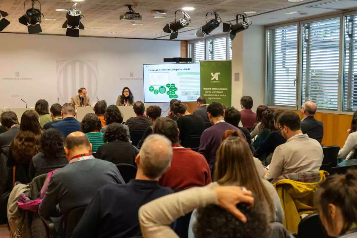 Una conferencia en una sala con varias personas sentadas escuchando a dos presentadores frente a una pantalla que muestra una presentación.