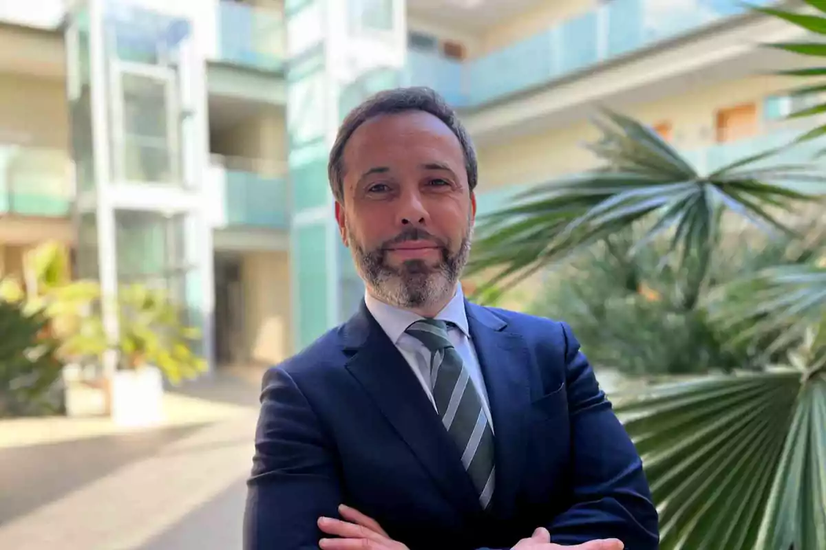 Un hombre con traje y corbata posa con los brazos cruzados en un entorno moderno con plantas y arquitectura contemporánea.