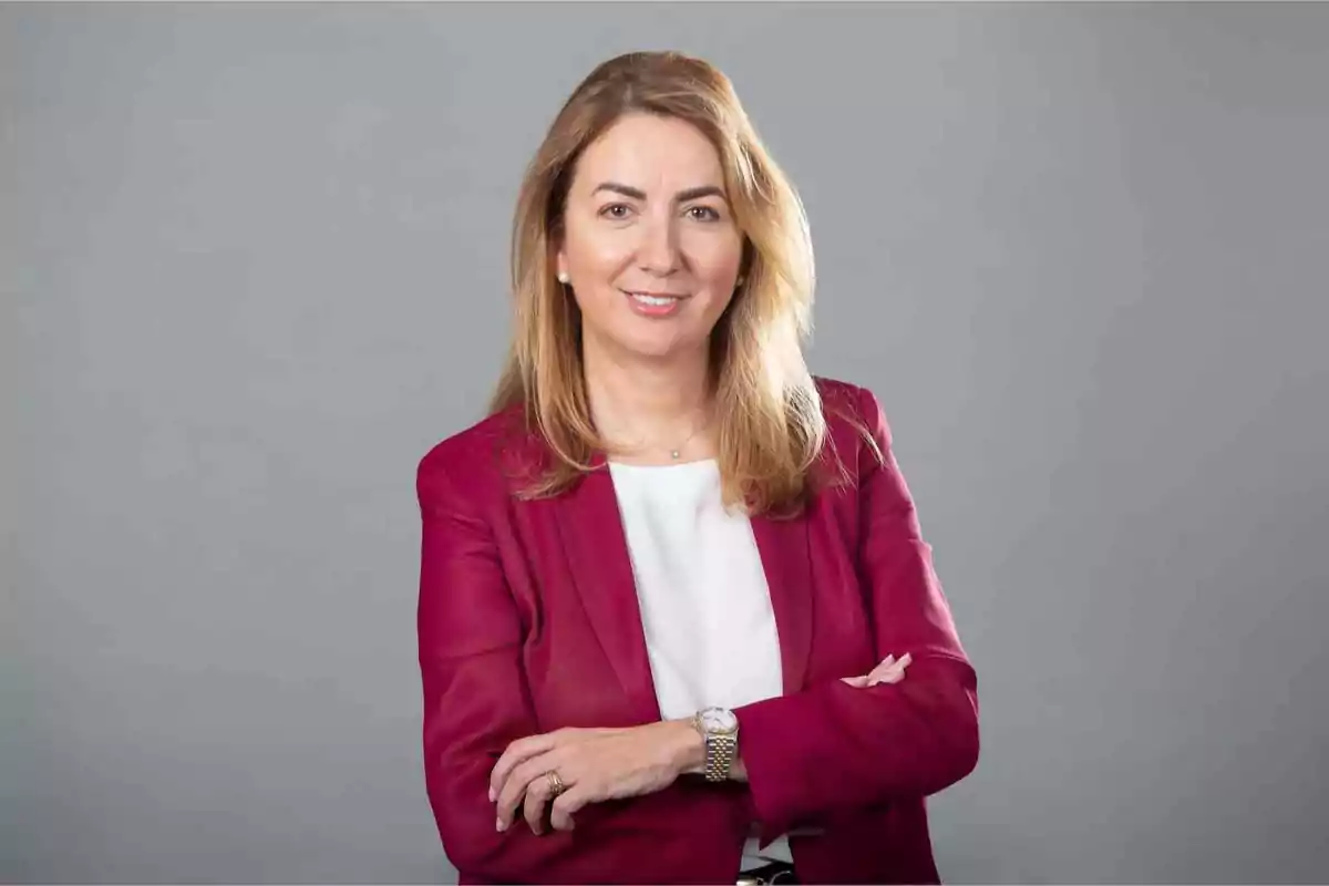 Mujer con chaqueta rosa y camisa blanca sonriendo con los brazos cruzados frente a un fondo gris.