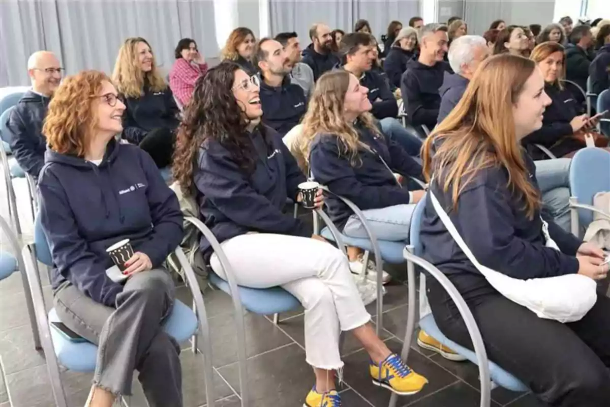 Un grupo de personas sentadas en sillas azules, vistiendo sudaderas oscuras, sonríen y parecen estar disfrutando de una presentación o evento en un espacio cerrado.